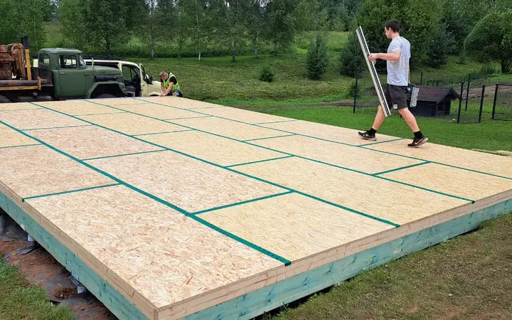 Ieklāts OSB grīdas klājs uz pamatiem, šuves noblīvētas ar lentu; strādnieks nes materiālu.
