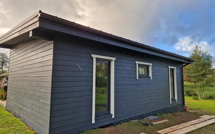 Pabeigta vienstāva karkasa māja ar tumši pelēku koka apšuvumu un augstiem logiem baltās ailēs.